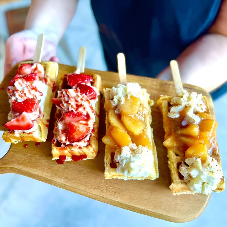 Four waffle pops, two with strawberries and whipped cream, two with peaches and whipped cream.