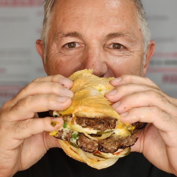 A man with gray hair takes a large bite of a Pudgee's double cheeseburger with onions and pickles.